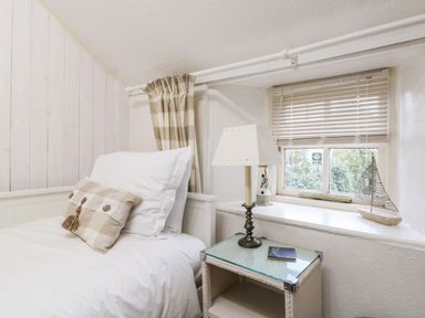 A bedroom with a white bed, checkered pillow, bedside table with a lamp and window with blinds at Pippin in Lelant