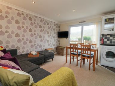 A living room with a grey sofa purple cushions a wooden dining table and chairs a wall mounted tv and sliding glass doors at Ocean Blue in Porthtowan