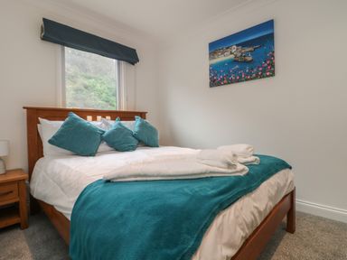 A bedroom with a wooden bed with white bedding and teal pillows and blanket with a window and a painting on the wall at Ocean Blue in Porthtowan