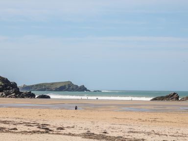 A sandy beach with some rocks on either side a person near the shore and small groups of people walking along the water at Bayview in Watergate Bay