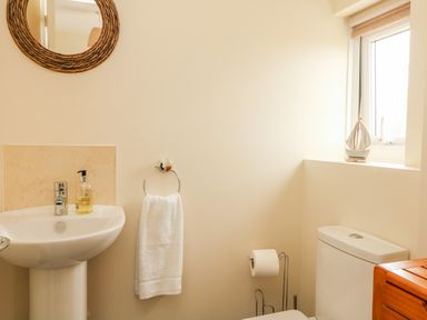 A bathroom with a sink a round mirror a towel holder with a white towel a toilet paper holder a window and a small sailboat decoration at Bayview in Watergate Bay