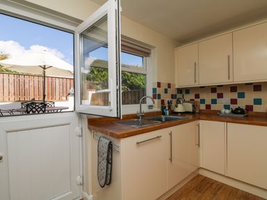 A kitchen with wooden countertops cream cabinets a sink and an open door leading to an outdoor patio with a table and umbrella at Bayview in Watergate Bay
