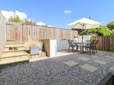 An outdoor patio area with a table and chairs under a large umbrella and stone steps with a small dog lying on them at Bayview Watergate Bay