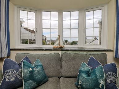 A sofa with blue and green cushions under a bay window with a small sailboat decoration at Bayview in Watergate Bay