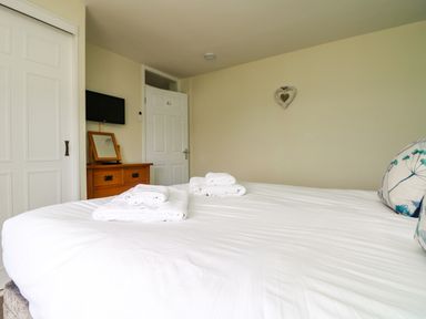 A bedroom with a bed with white bedding and folded towels a wooden dresser a wall-mounted television and a white door at Bayview Watergate Bay