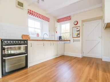 A kitchen with wooden floor white cabinets black oven two windows with red and white checkered curtains and a white door at Goshen Farm in St Agnes