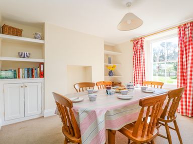 A dining room with wooden chairs and a table set for tea by a window with red check curtains at Goshen Farm in St Agnes