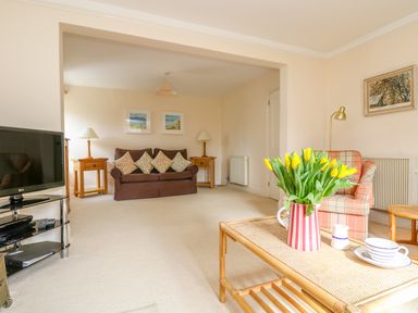 A living room with a brown sofa with cushions two wooden side tables with lamps a plaid armchair a coffee table with yellow flowers and tea set a tv on a stand and paintings on the walls at Goshen Farm in St Agnes