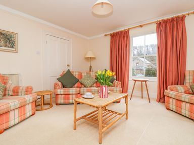 A living room with plaid sofas, a wooden coffee table with flowers, a window with curtains, and a floor lamp at Goshen Farm in St Agnes