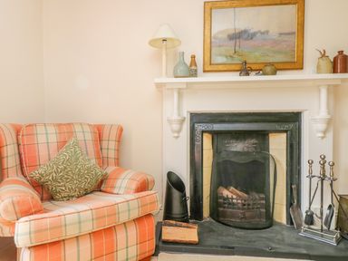 A living room with an armchair beside a fireplace with fire tools and decorative items at Goshen Farm in St Agnes