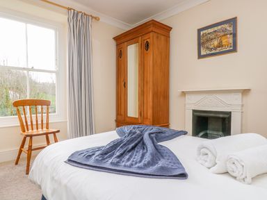 A bedroom with a wooden chair by a window, a wooden wardrobe with a mirror, a fireplace, a bed with a blue blanket and two white rolled towels at Goshen Farm in St Agnes