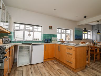 A kitchen with wooden cabinets a small fridge double windows and a dining table at Trelawns in Mawgan Porth