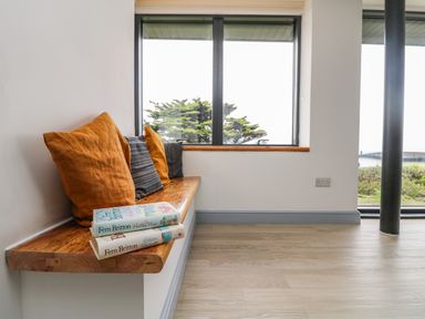 A wooden bench with cushions and two books under a window with a view of trees at Trelawns in Mawgan Porth