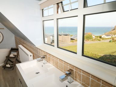 A bathroom with a double sink, taps, tiled wall, window with ocean view and a bathtub at Trelawns in Mawgan Porth