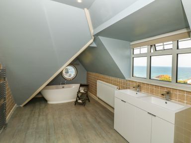 A bathroom with a bathtub under a sloped ceiling and a double sink vanity by a large window overlooking the sea at Trelawns in Mawgan Porth