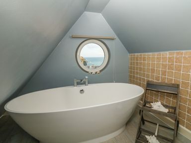 A bathroom with a freestanding bathtub a round window showing an ocean view and a wooden chair with shells at Trelawns in Mawgan Porth
