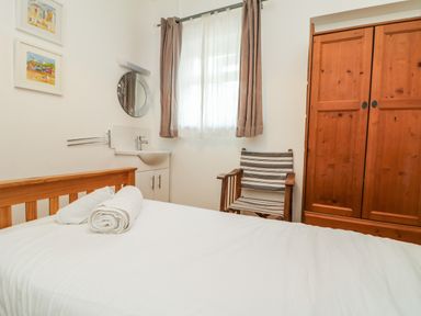 A bedroom with a bed with white sheets and rolled towels a wooden wardrobe a striped chair a round mirror and a window with curtains at Trelawns in Mawgan Porth