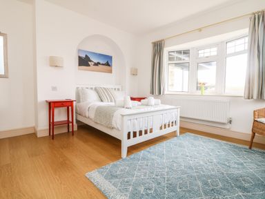 A bedroom with a white bed a blue rug red bedside tables and a window with curtains at Trelawns in Mawgan Porth