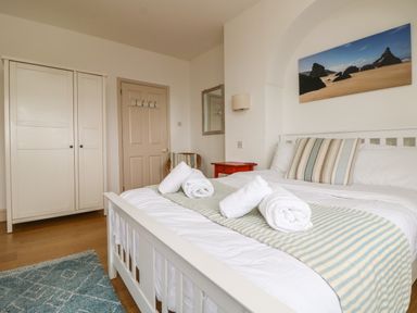 A bedroom with a white bed with rolled towels on it a wardrobe a door a red bedside table and a picture of a beach at Trelawns in Mawgan Porth