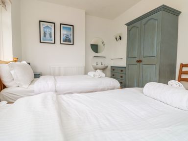 A bedroom with two single beds white linens a gray wardrobe a dresser a wall-mounted sink and framed pictures at Trelawns in Mawgan Porth