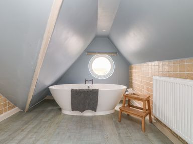 A bathroom with a white freestanding bathtub under a sloped ceiling and a round window at Trelawns in Mawgan Porth