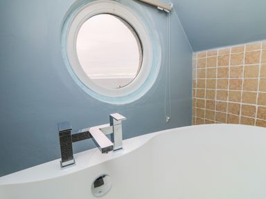 A bathroom with a white bathtub and a round window with a view of the sea at Trelawns in Mawgan Porth