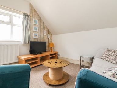 A small living room with a wooden TV stand a round wooden spool coffee table a gray sofa with striped cushions a blue armchair and a window at Trelawns in Mawgan Porth
