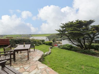 An outdoor patio with wooden benches and a table on a stone surface overlooking a grassy garden with a tree and distant houses at Trelawns in Mawgan Porth