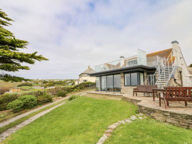 A house with a glass railing balcony wooden benches and a stone path on a grassy area at Trelawns in Mawgan Porth