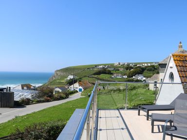 A balcony with outdoor chairs and a table overlooking a coastal village and the ocean at Trelawns in Mawgan Porth