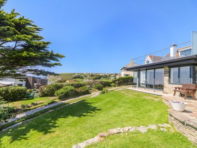 A grassy garden with shrubs and a stone path next to a house with large windows at Trelawns in Mawgan Porth