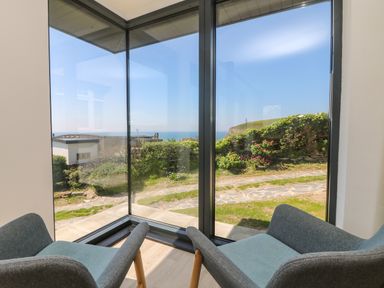 Corner of a room with two chairs facing large windows overlooking a garden path and ocean at Trelawns in Mawgan Porth