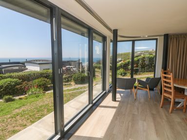 A room with large glass windows two gray chairs a wooden table and chairs overlooking a garden and ocean view at Trelawns in Mawgan Porth