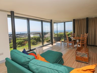 A living room with green sofa wooden chairs round table and large windows showing a garden and ocean view at Trelawns in Mawgan Porth
