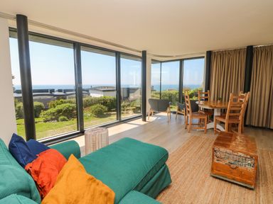 A living room with teal sofa, wooden dining table with chairs, armchairs, wooden chest, and large windows with garden and sea view at Trelawns in Mawgan Porth