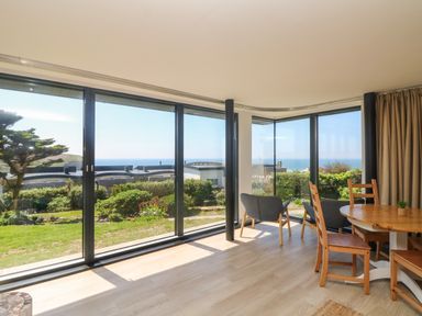A room with large glass windows showing a garden and ocean view with two grey armchairs and a wooden dining table with chairs at Trelawns in Mawgan Porth
