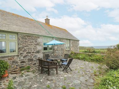 An outdoor patio with a wooden table and chairs with a blue umbrella next to a stone house with green windows at Parc An Castle Cottage in Lizard