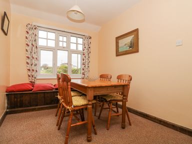 A dining room with wooden table and four chairs next to a window with curtains and a window seat with cushions at Parc An Castle Cottage in Lizard