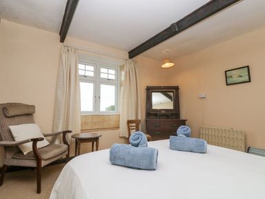 A bedroom with a bed, two rolled blue towels on the bed, a cushioned armchair, a small round wooden table, a wooden dresser with a mirror, and a window with curtains at Parc An Castle Cottage in Lizard