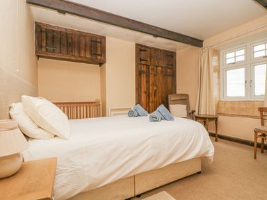 A bedroom with a bed, bedside table with lamp, wooden cupboards, chair, window with curtains at Parc An Castle Cottage in Lizard
