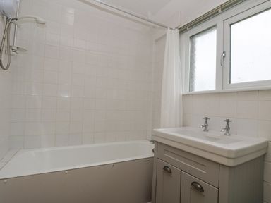 A bathroom with a bathtub shower combo white tiles a sink cabinet and a frosted window at Parc An Castle Cottage in Lizard
