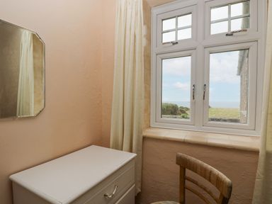 A small room with a white chest of drawers a wooden chair a window with a view of the outdoors and a mirror on the wall at Parc An Castle Cottage in Lizard