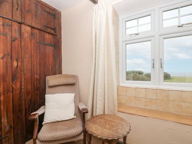 A room corner with a patterned armchair with a white pillow next to a small wooden table and a window showing bushes and sky at Parc An Castle Cottage in Lizard
