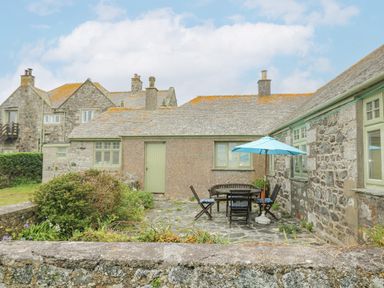 An outdoor patio with wooden chairs and a table under a blue umbrella beside stone cottages at Parc An Castle Cottage in Lizard
