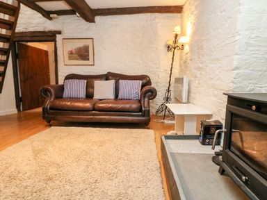 A living room with a brown leather sofa with cushions a wooden floor a beige rug a white side table a floor lamp and a wood burning stove at Apple Loft in Lostwithiel