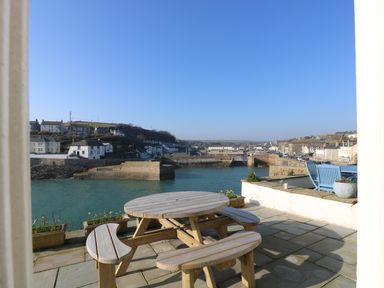 A patio with a round wooden picnic table and flower pots overlooking a harbor with buildings in the background at Haven in Porthleven