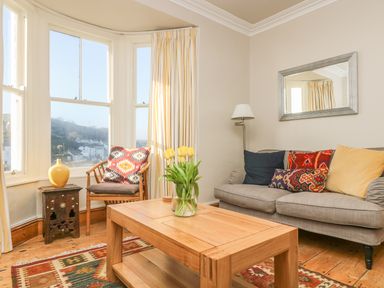 A living room with a gray sofa with colorful cushions a wooden coffee table with a vase of yellow tulips a wooden chair with a patterned cushion a small wooden side table with a yellow vase a floor lamp a wall mirror and large windows with curtains at Haven in Porthleven