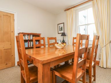A dining room with a wooden table and six chairs a bookshelf a lamp a window with striped curtains and a wooden door at Haven in Porthleven