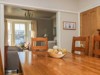 A wooden dining table with decorative balls in a bowl and wooden chairs in a dining room with a view into a living room with a sofa and a bay window at Haven in Porthleven
