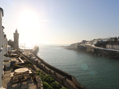 A coastal view with a stone walkway, parked cars, houses on cliffs, and a clock tower at Haven in Porthleven